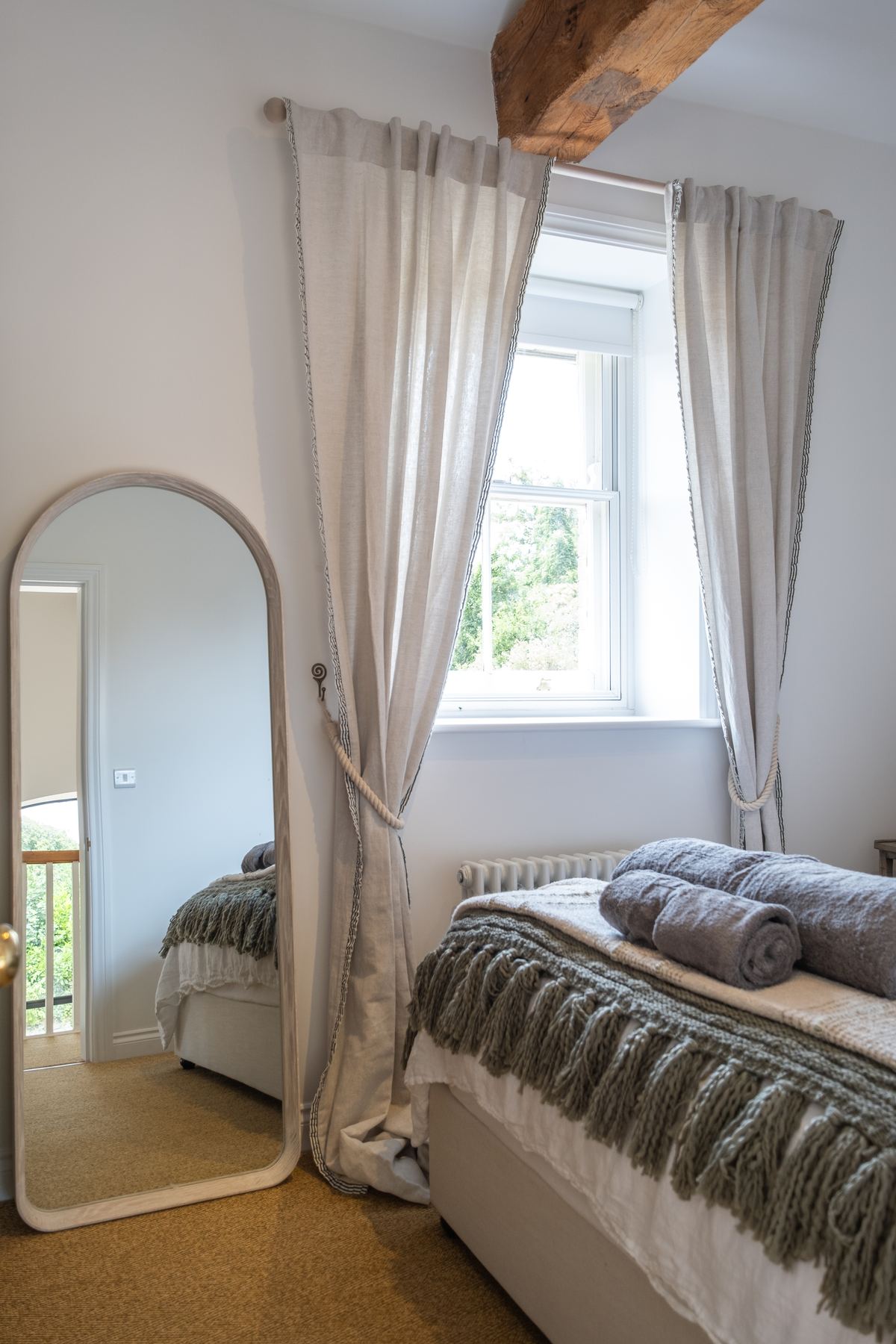 A bedroom with an arched floor mirror and linen curtains beneath an old beam