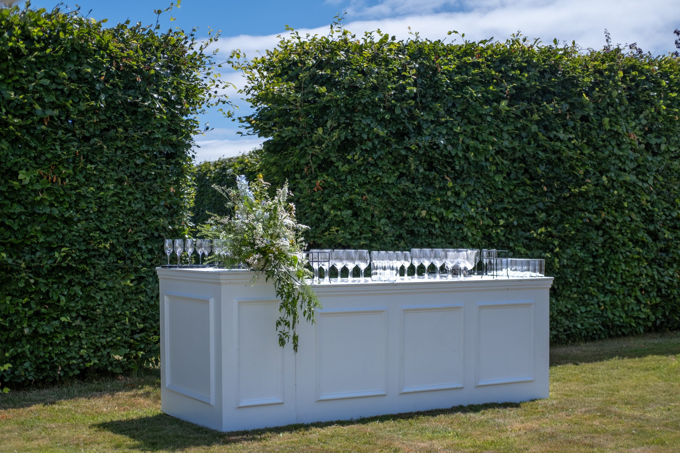 A white panelled bar dressed with glasses and wild flowers, against a tall beech hedge
