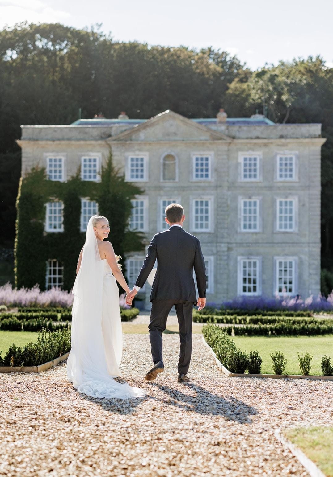 A bride and groom walking hand in hand up the gravel towards Gatcombe House