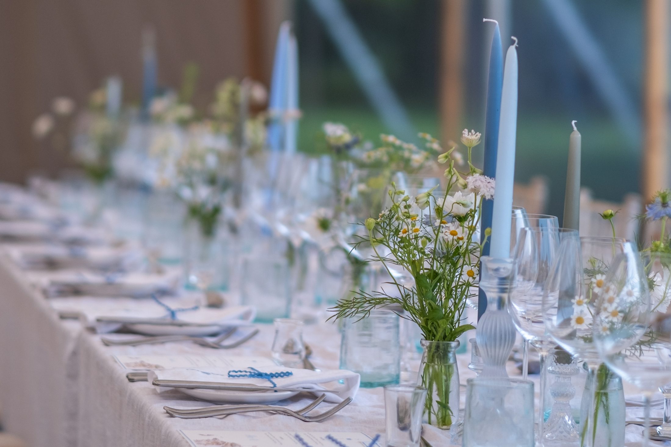 Place settings with blue ribbon, tapered candles and jars of wild flowers along a long table